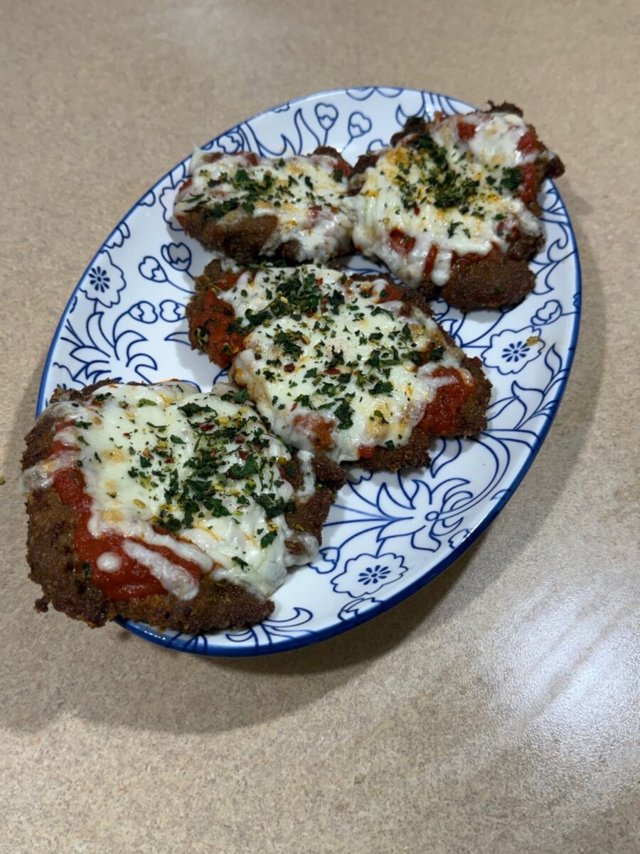 Four pieces of breaded chicken topped with marinara sauce, melted cheese, and herbs are arranged on a white and blue floral-patterned plate, set on a light brown countertop.