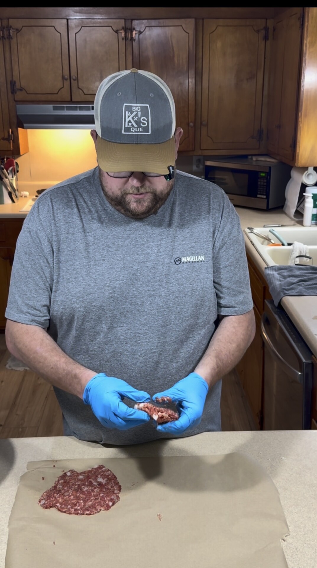 A man wearing a gray shirt, cap, and blue gloves forms ground meat into a patty over parchment paper in a kitchen with wooden cabinets.
