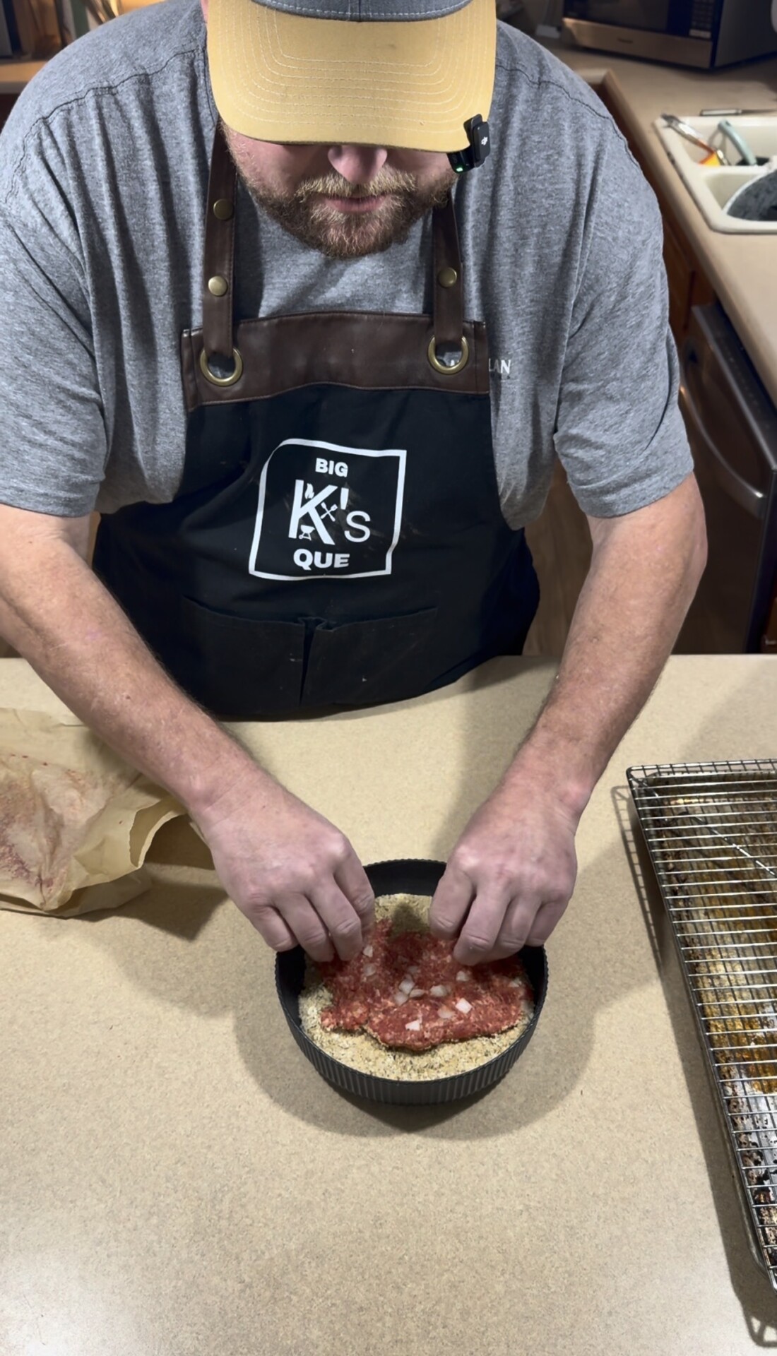A person wearing a dark apron and a gray shirt presses ground meat into a round pan on a kitchen counter, preparing food. A wire rack and some parchment paper are nearby.