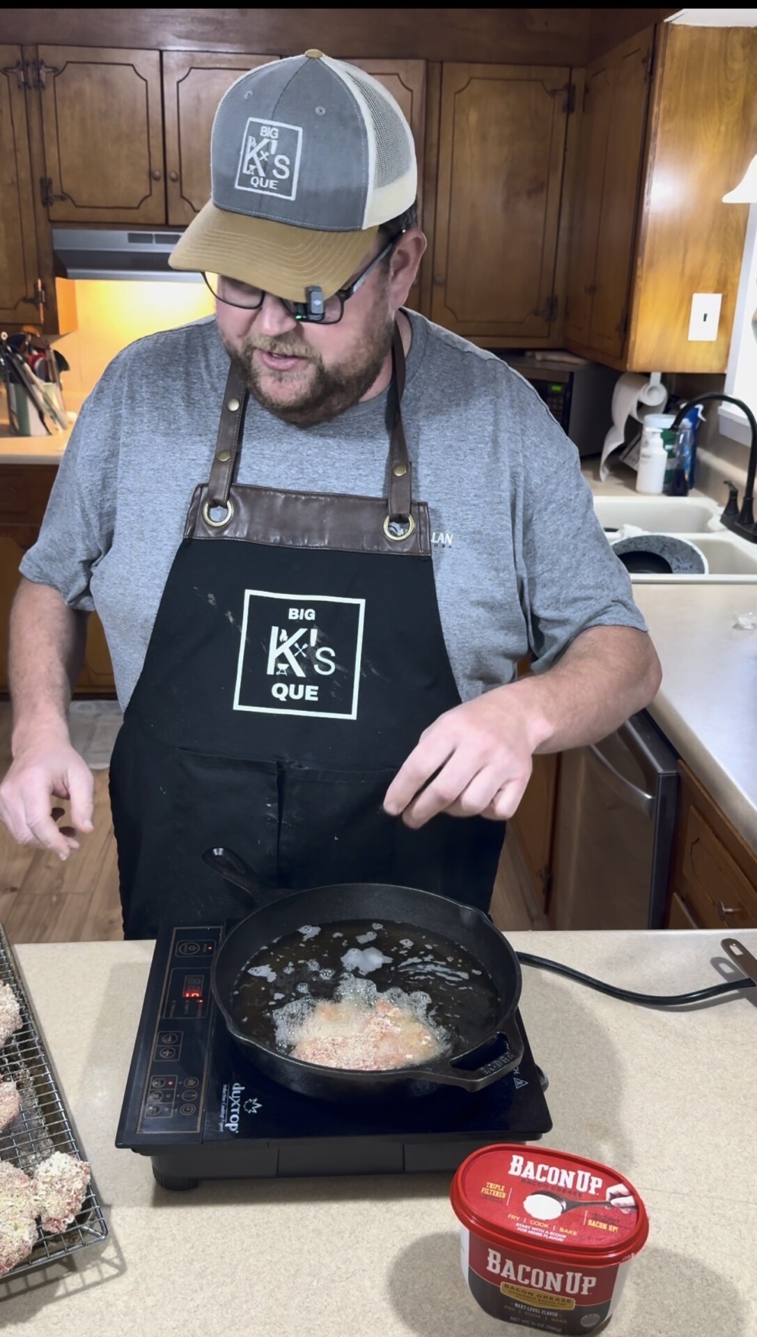A man wearing a hat and apron fries food in a skillet on an electric cooktop in a kitchen. A container labeled Bacon Up sits nearby. Breaded items rest on a rack next to him.