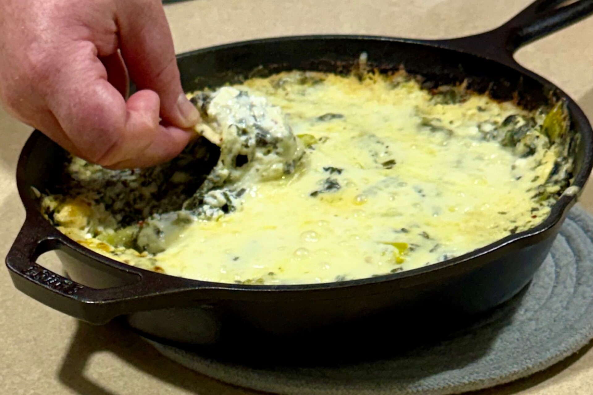 A hand dips a chip into a cast iron skillet filled with creamy, baked spinach and cheese dip on a stove.