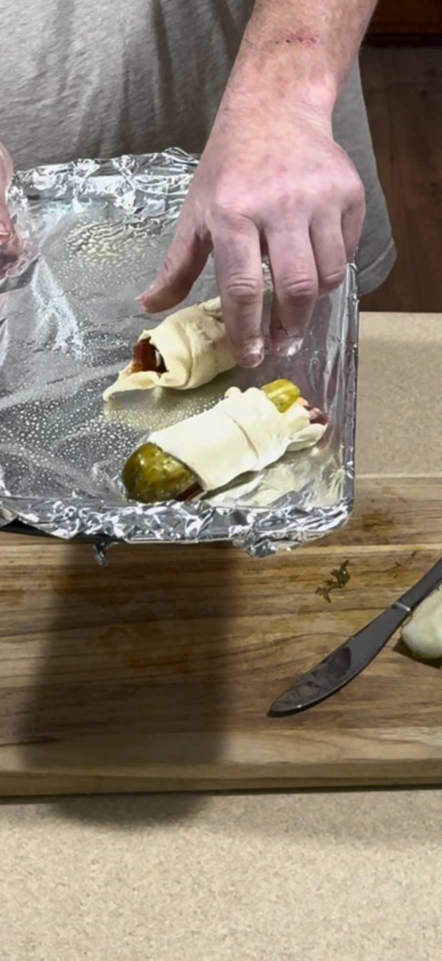 A person holds a foil-lined tray with two pickles wrapped in pastry dough and sliced meat. A knife and sliced onion sit on a wooden cutting board nearby.