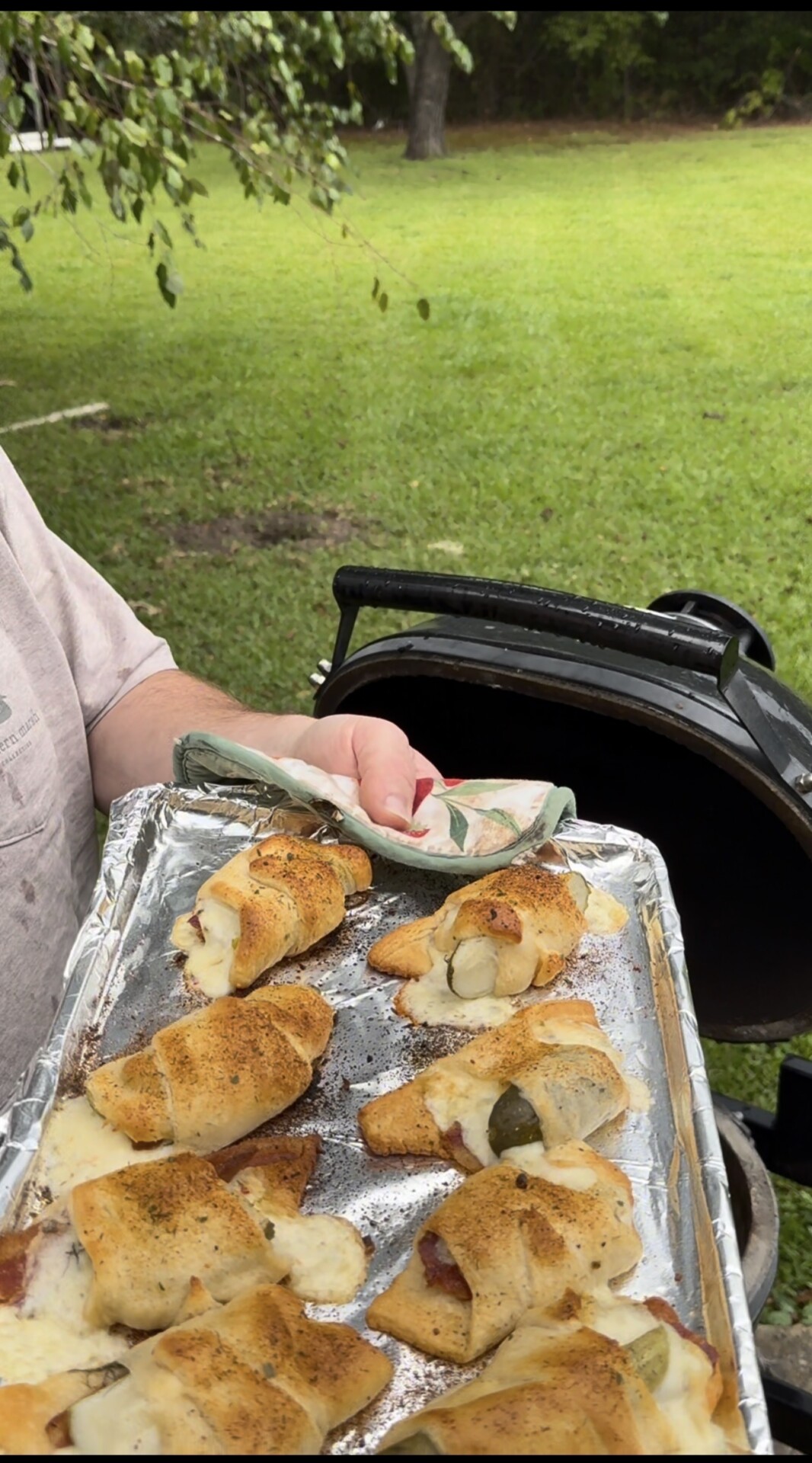A person holds a foil-lined tray of baked crescent rolls with melted cheese and seasoning, standing next to a grill in a green backyard.
