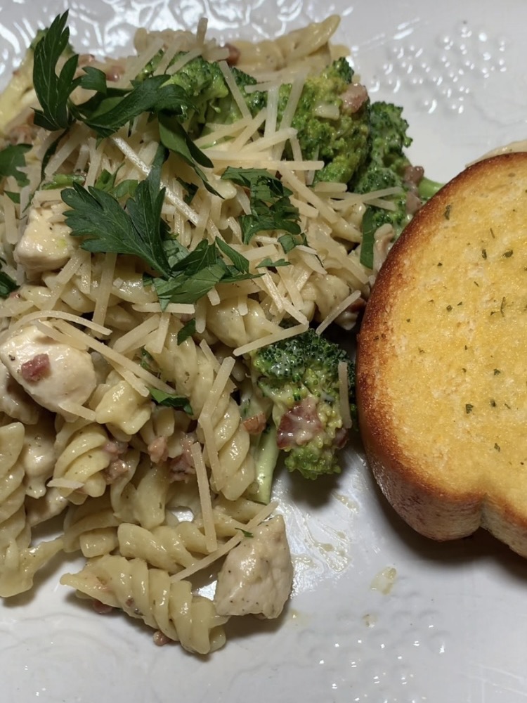 A plate of rotini pasta with chicken, broccoli, bacon bits, shredded parmesan, and parsley, served with a slice of garlic bread on a white plate.
