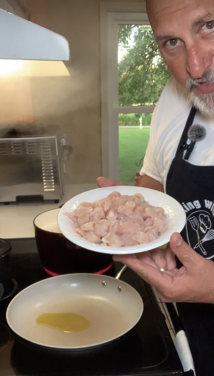 A man in a kitchen holds a plate of raw, diced chicken over a stove with a pan of olive oil, ready to cook. There is a window with greenery outside in the background.