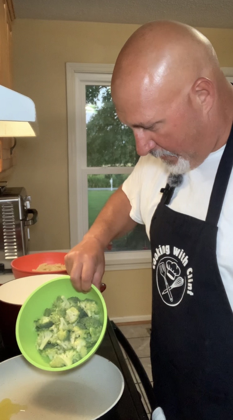 A man in a Cooking With Clint apron pours broccoli from a green bowl into a pan on a stove in a kitchen, with a window showing trees in the background.