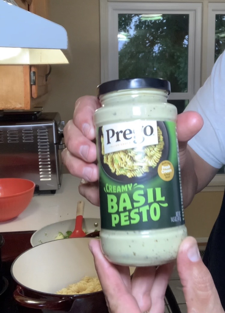 A person holds a jar of Prego Creamy Basil Pesto sauce in a kitchen. A stove, red bowl, and pot with pasta are visible in the background.