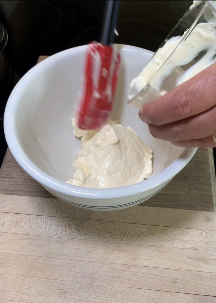 A person uses a red spatula to scrape a creamy mixture from a container into a white mixing bowl on a wooden surface.
