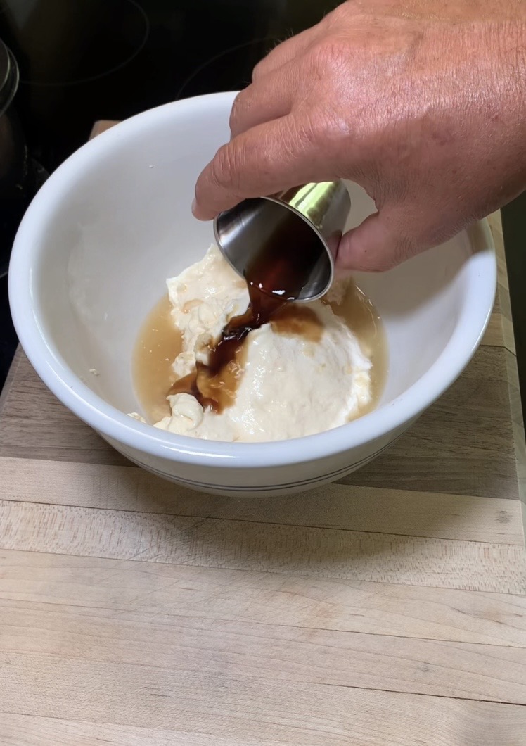 A hand pours a dark liquid from a small metal cup into a white bowl containing a creamy mixture and a light tan liquid, all set on a wooden surface.