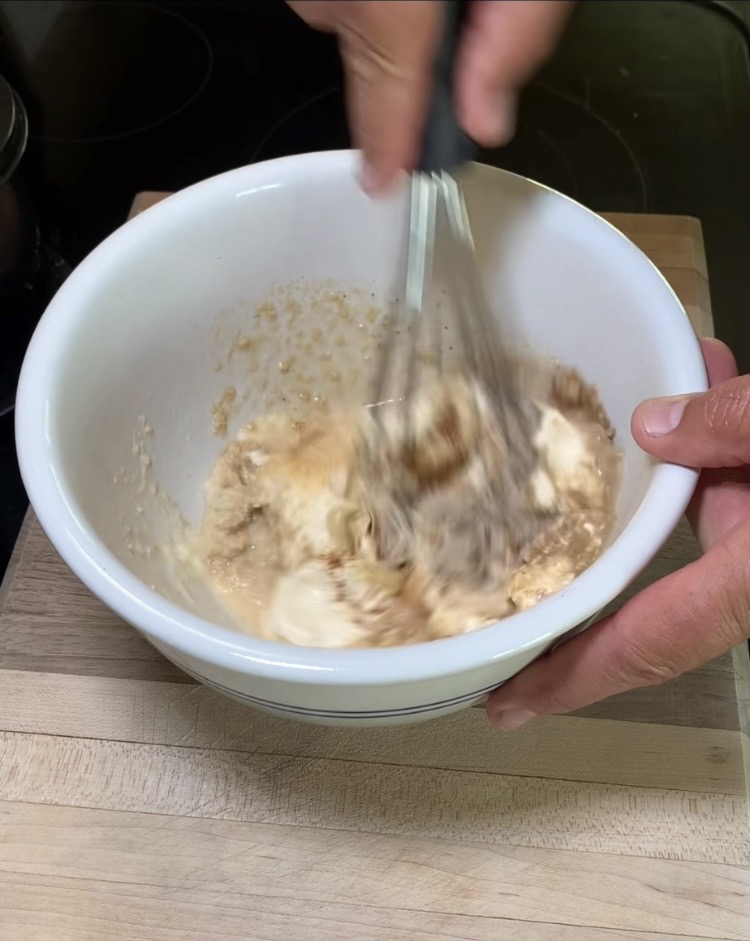 A person is whisking a mixture of creamy and brown ingredients in a white bowl placed on a wooden surface.