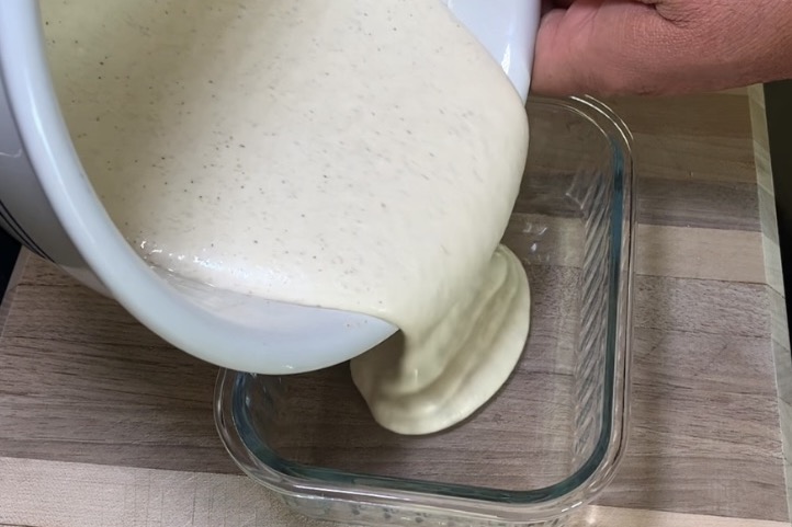 A person pours thick, creamy batter from a white bowl into a rectangular glass dish on a wooden surface.
