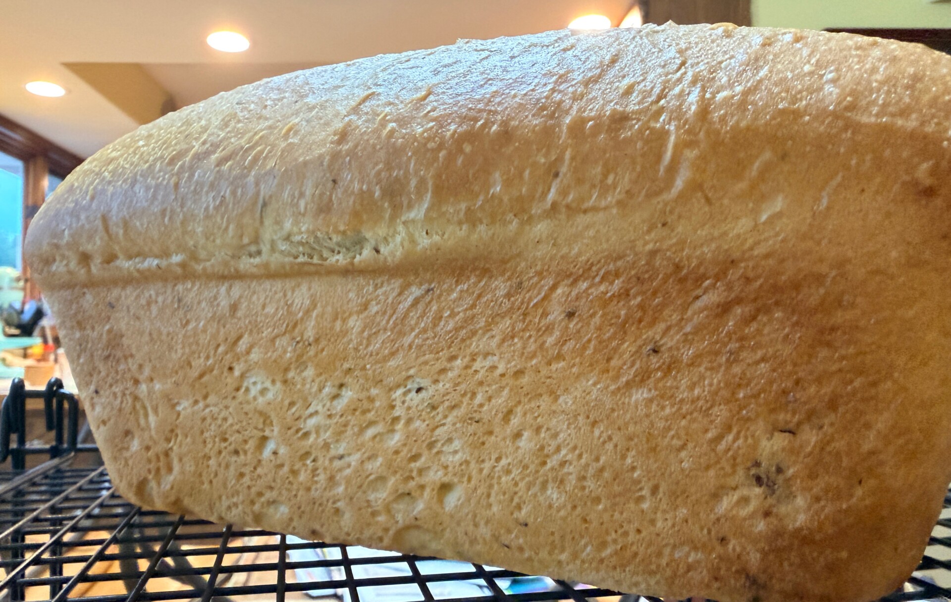 A close-up of a freshly baked loaf of bread cooling on a wire rack, with a golden-brown crust and airy texture. The kitchen background is softly out of focus.