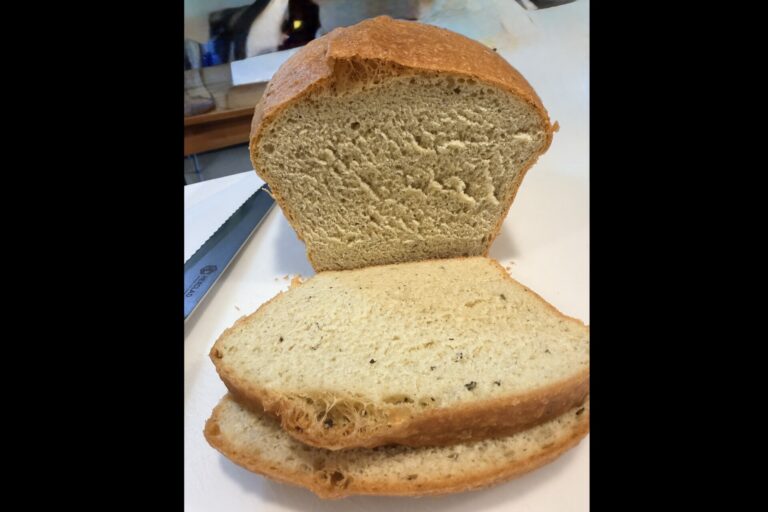A loaf of bread is sliced, with the top half standing upright and two slices lying flat in front. A bread knife is visible to the left on a white surface.