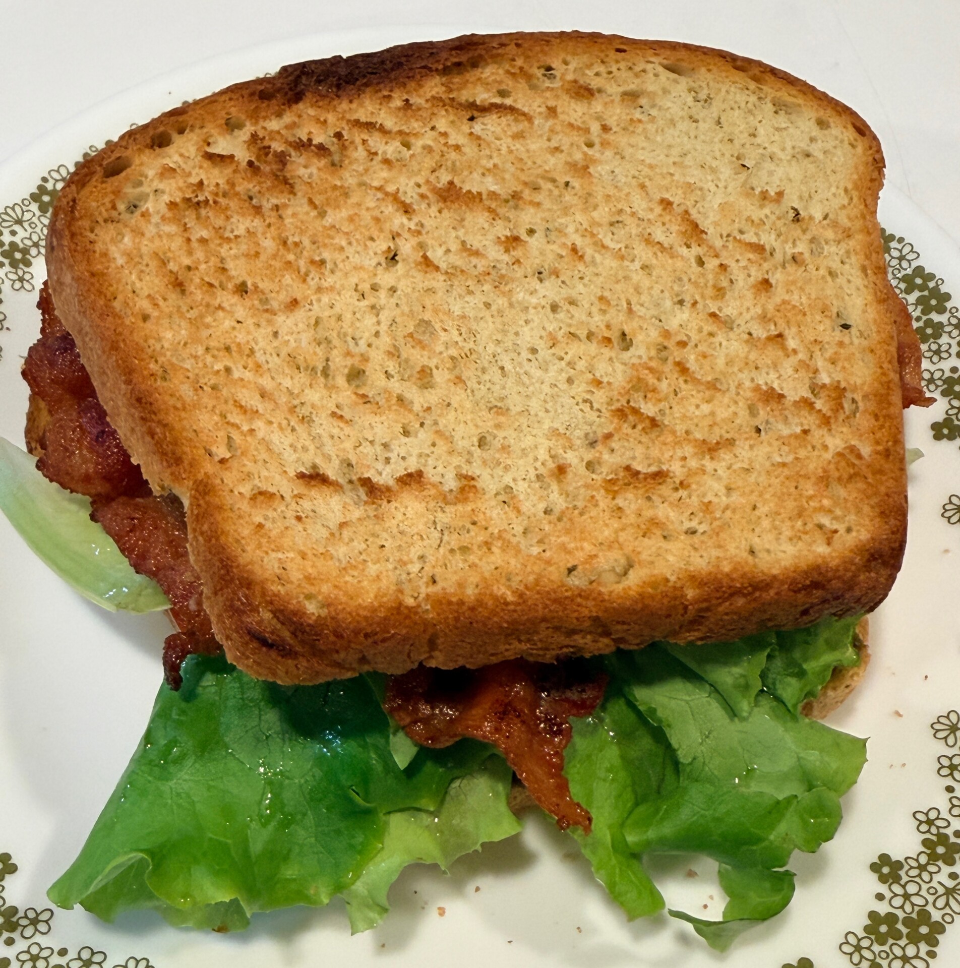 A BLT sandwich with lettuce and bacon peeking out from between two slices of toasted bread, served on a white plate with a green floral border.
