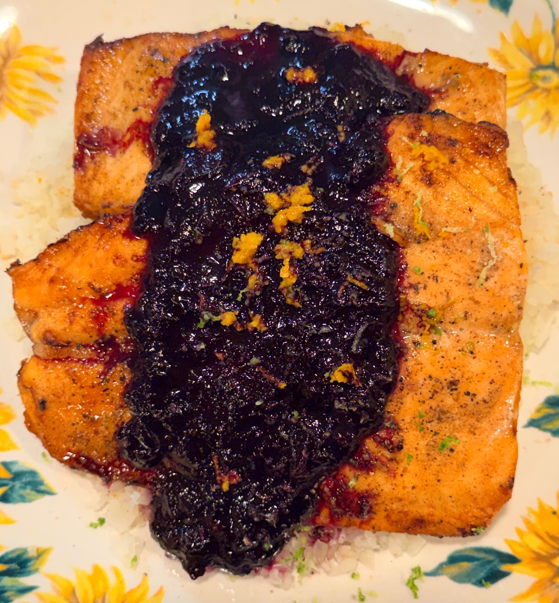Grilled salmon fillet topped with a dark berry sauce and orange zest, served over white rice on a floral-patterned plate.