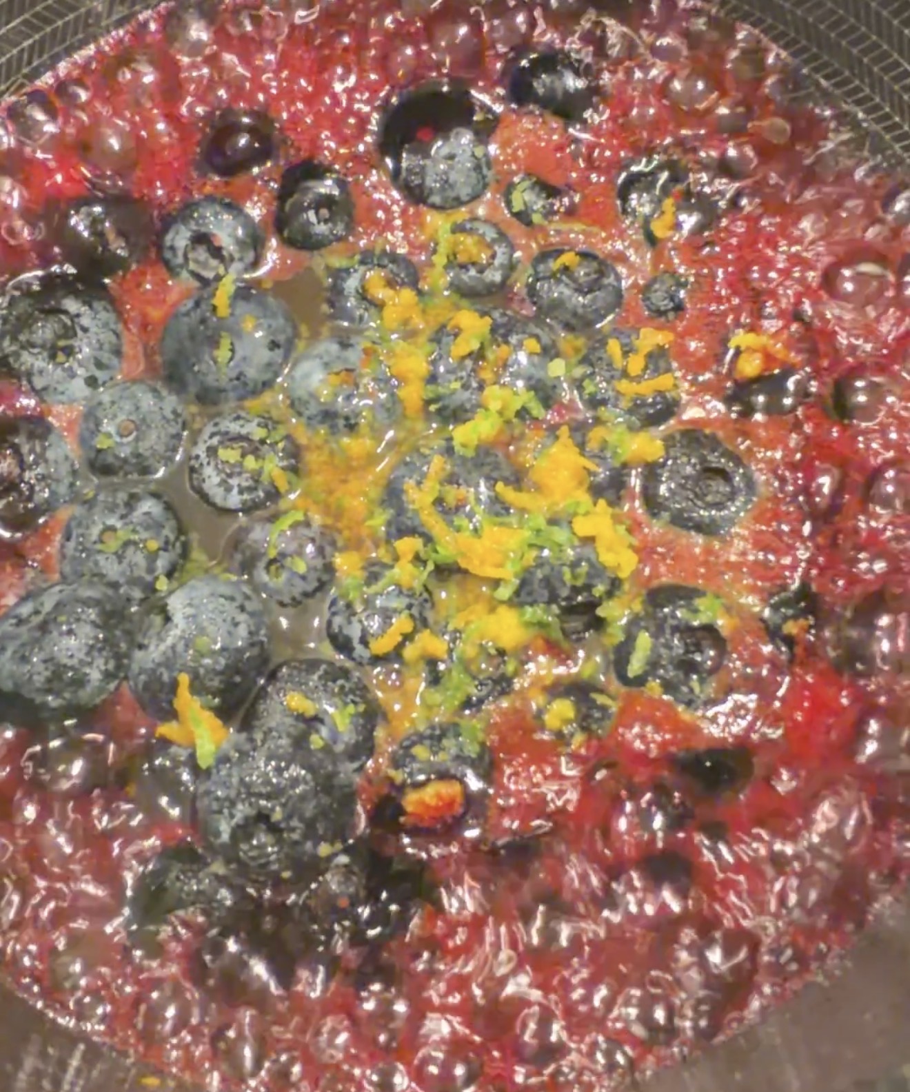 Blueberries and orange zest simmer in a bubbling red sauce, likely a fruit compote, inside a pan. The mixture appears thick and glossy, with vibrant colors and visible citrus zest on top.