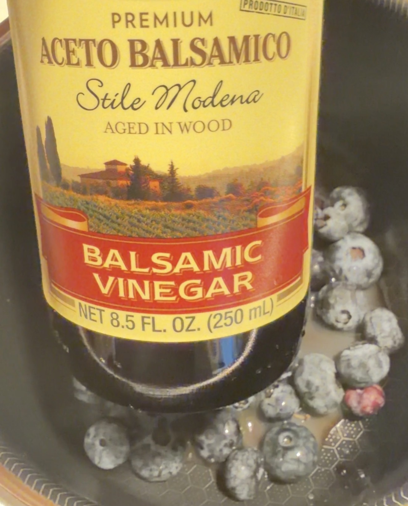 A bottle of balsamic vinegar is held over a pan with fresh blueberries and some liquid inside. The bottle label reads Aceto Balsamico Stile Modena..