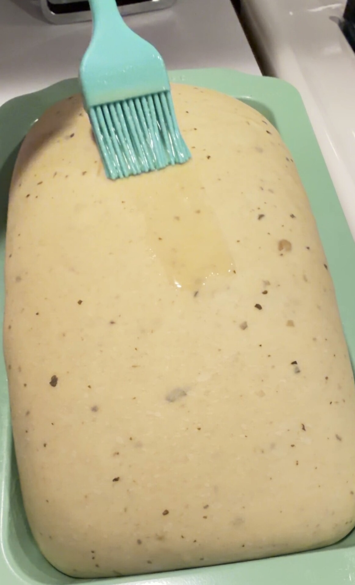 A blue brush spreads melted butter over an uncooked loaf of herb bread dough in a rectangular green pan.