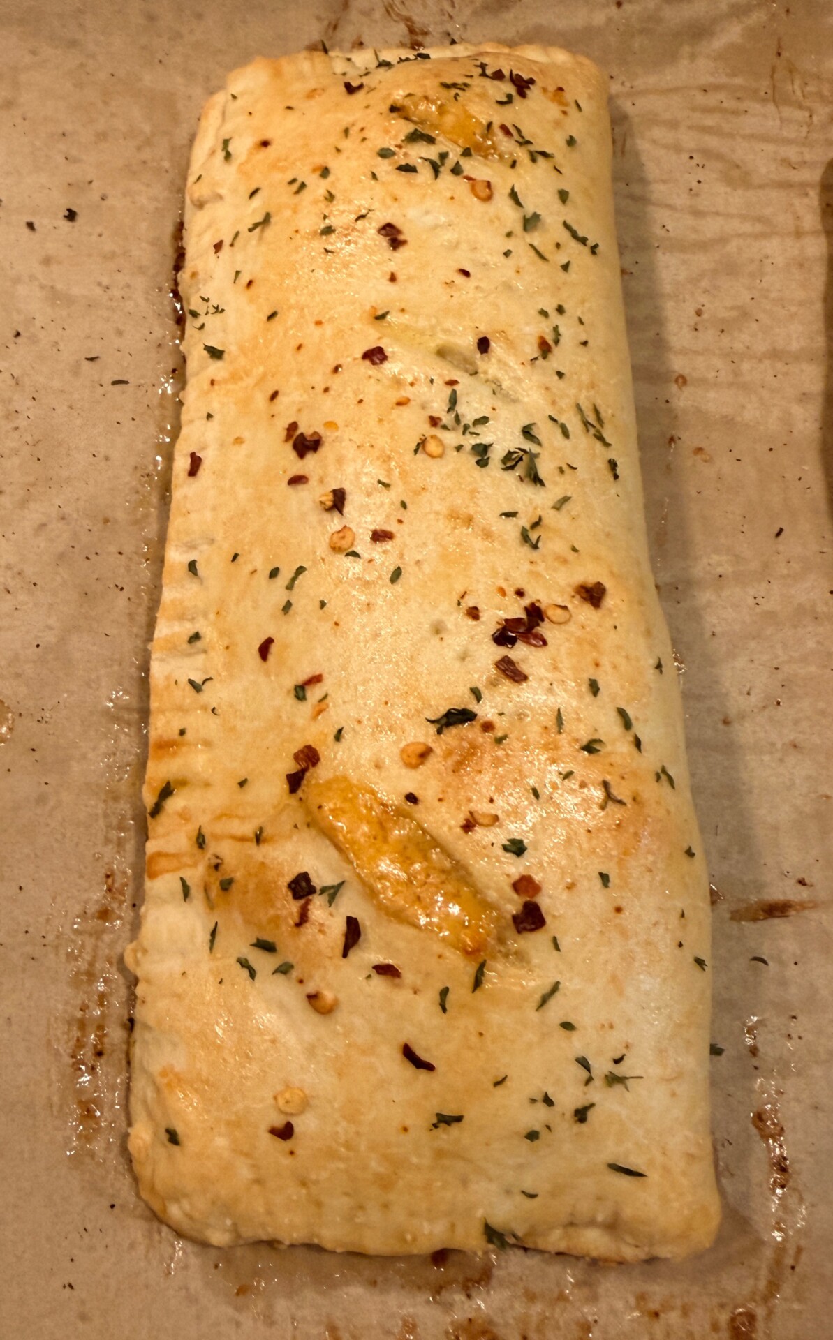 A baked, rectangular calzone or stuffed bread topped with herbs, red pepper flakes, and bits of melted cheese, resting on parchment paper.