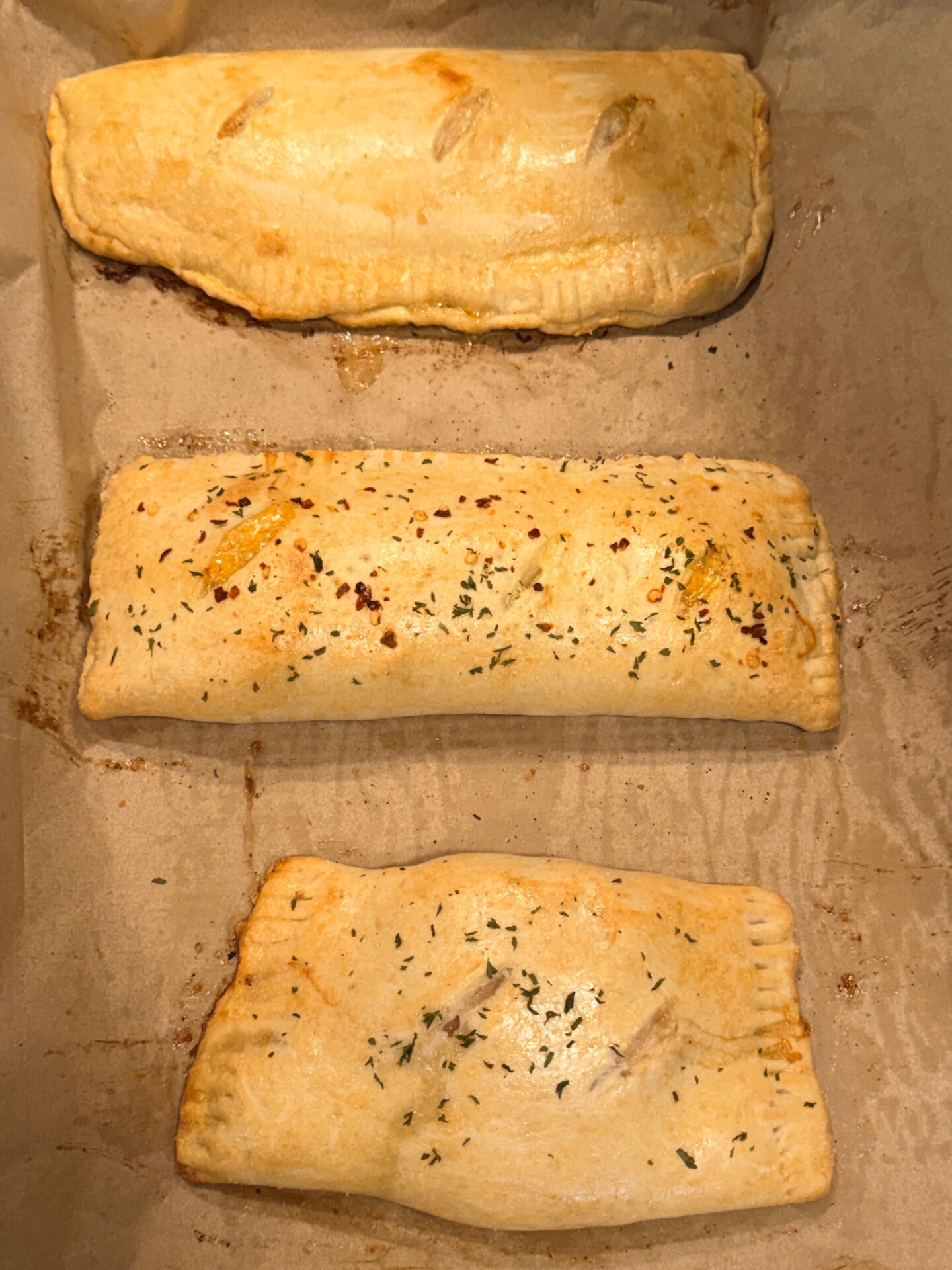 Three golden-brown pastry pockets with crimped edges are arranged on parchment paper, two topped with herbs and one plain and smooth.
