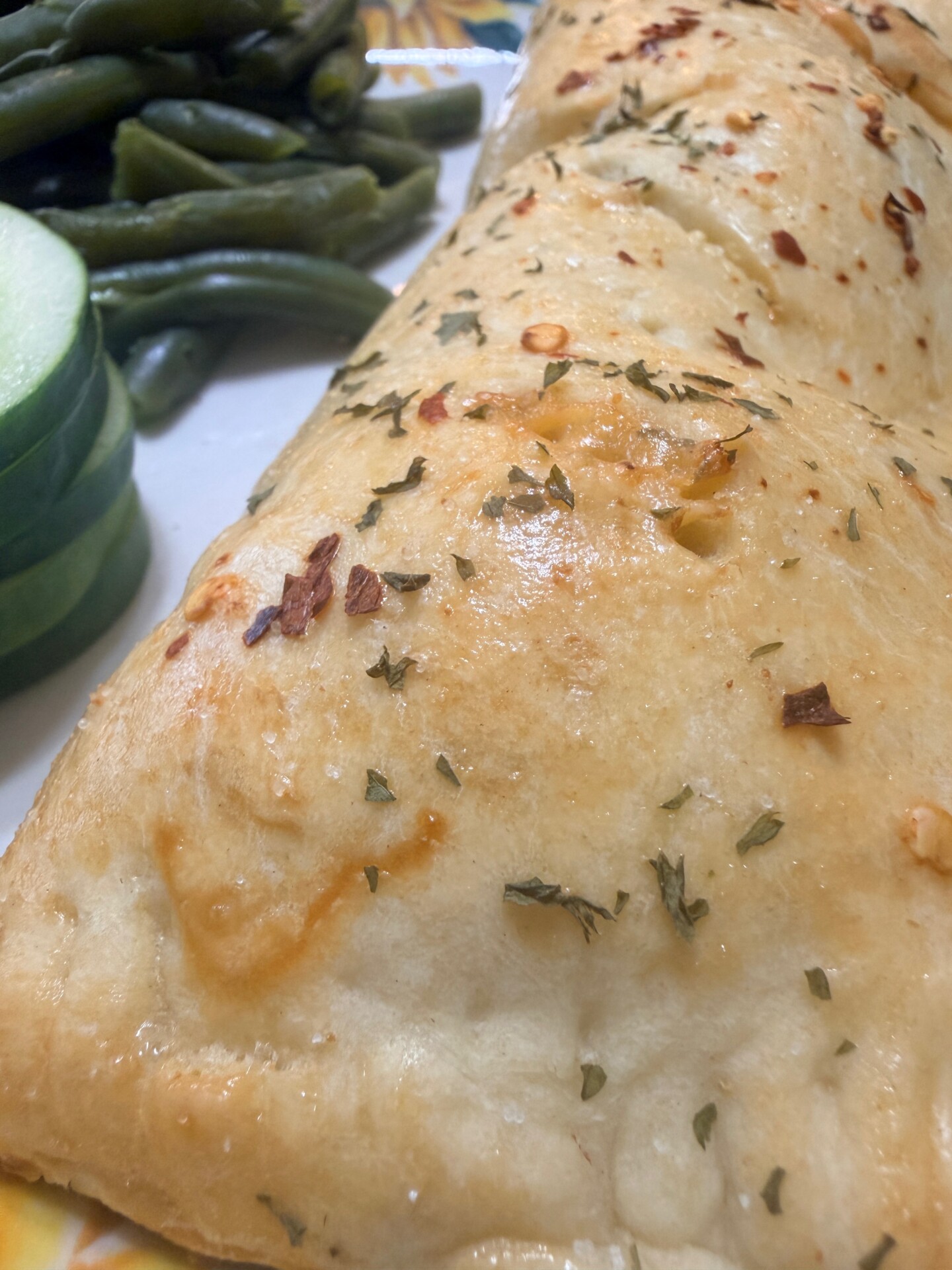 A golden-brown pastry topped with herbs sits on a plate next to green beans and cucumber slices.