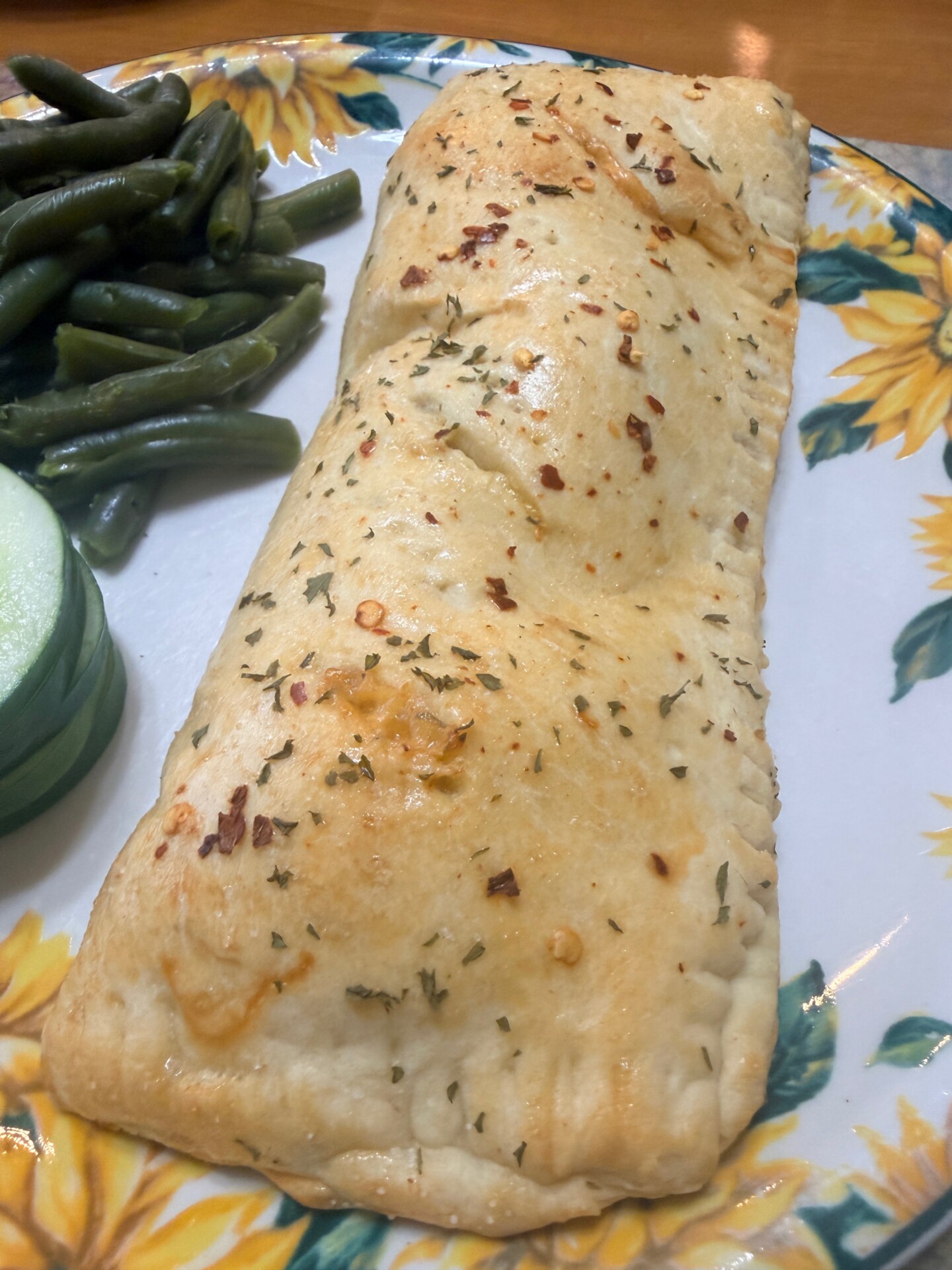 A golden-brown pastry pocket garnished with herbs on a sunflower-patterned plate, served with cut cucumber and green beans.