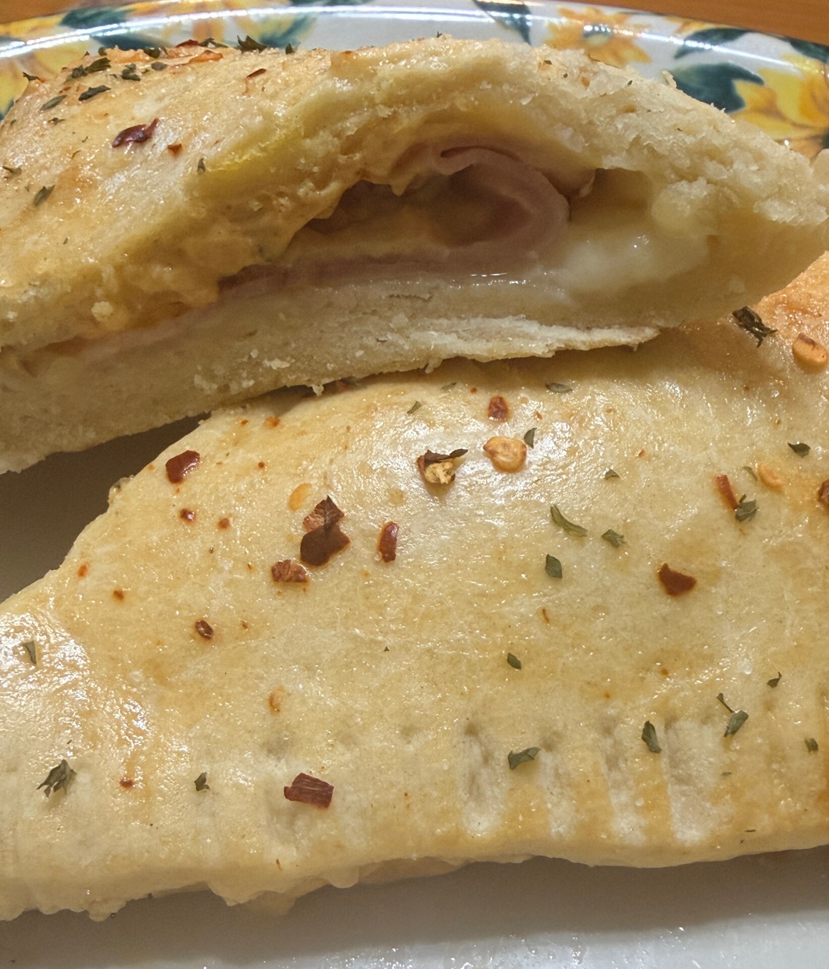 A golden-brown calzone cut in half, showing melted cheese and ham inside, topped with herbs and red pepper flakes, served on a floral plate.
