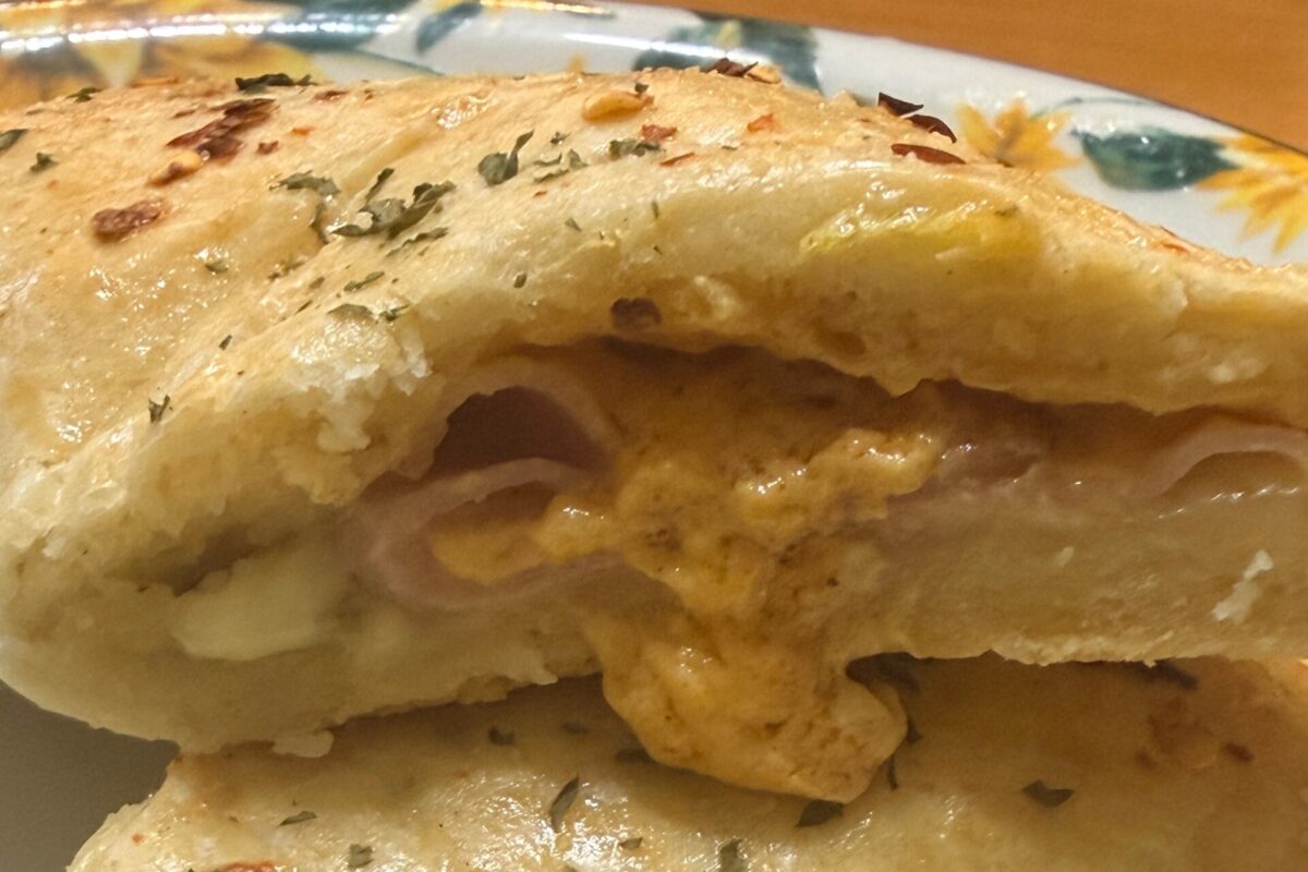 A close-up of a stuffed bread roll cut open, revealing melted cheese and sliced meat inside. The bread looks golden with herbs sprinkled on top, served on a floral-patterned plate.