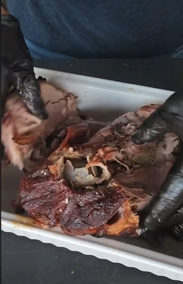 Person wearing black gloves is pulling apart a cooked, browned piece of meat in a white rectangular dish, revealing its tender, juicy inside.