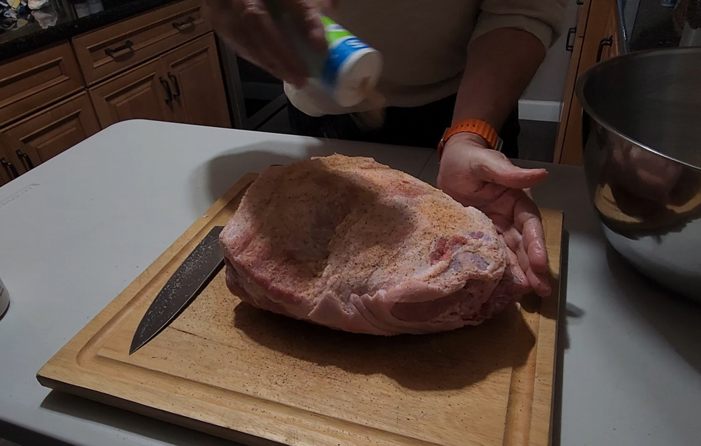 A person sprinkles seasoning from a container onto a large piece of raw meat on a wooden cutting board, with a knife and metal bowl nearby on the kitchen counter.