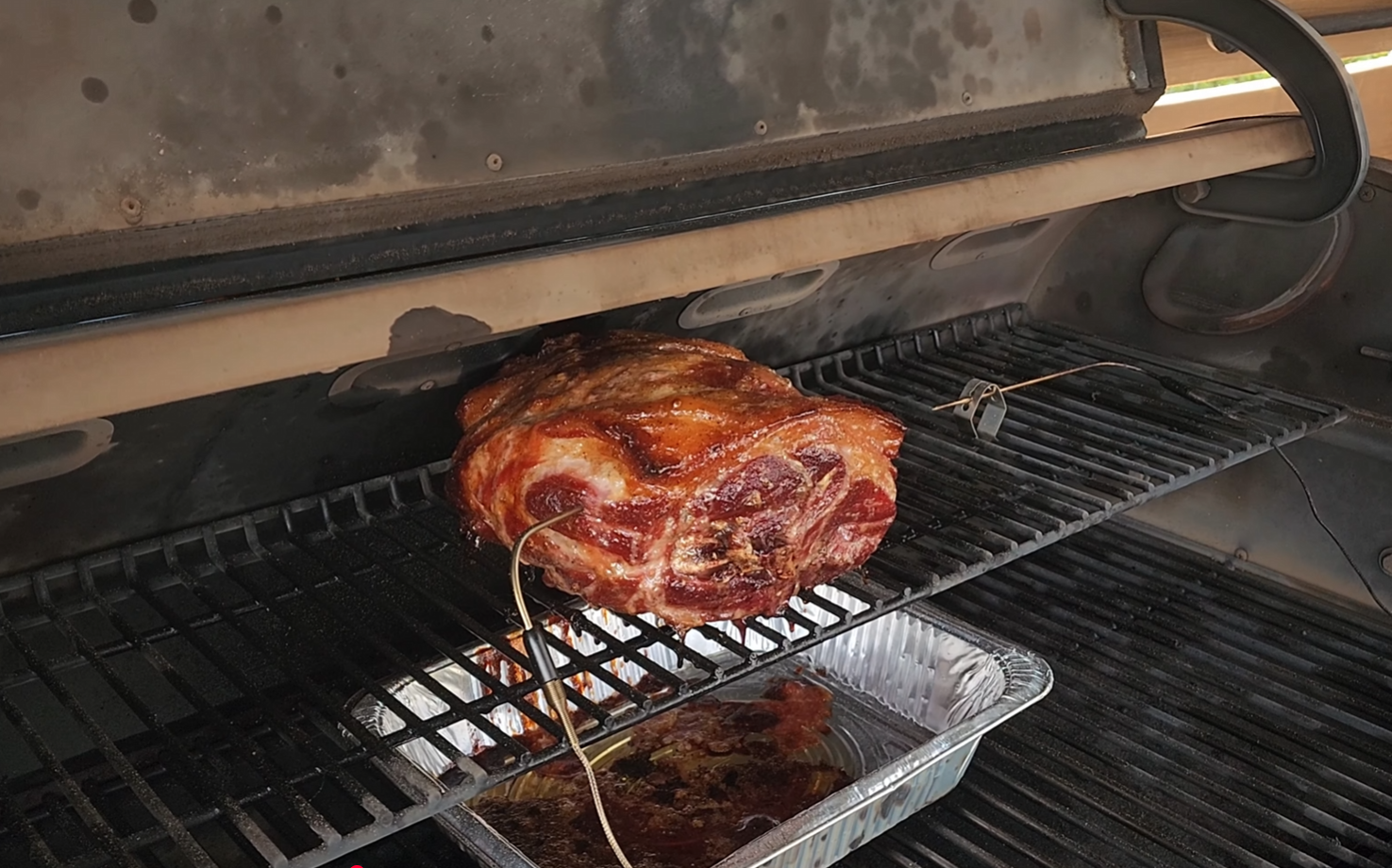 A large piece of meat is being smoked on a grill with two temperature probes inserted, and a foil tray underneath catching juices.