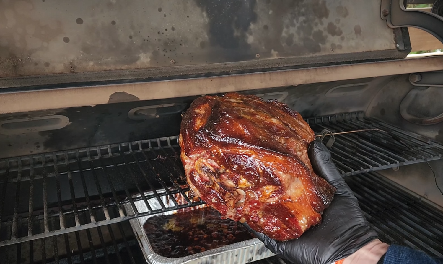 A gloved hand holds a glazed, smoked pork roast above a grill rack, with a foil pan underneath catching drippings inside a smoker.