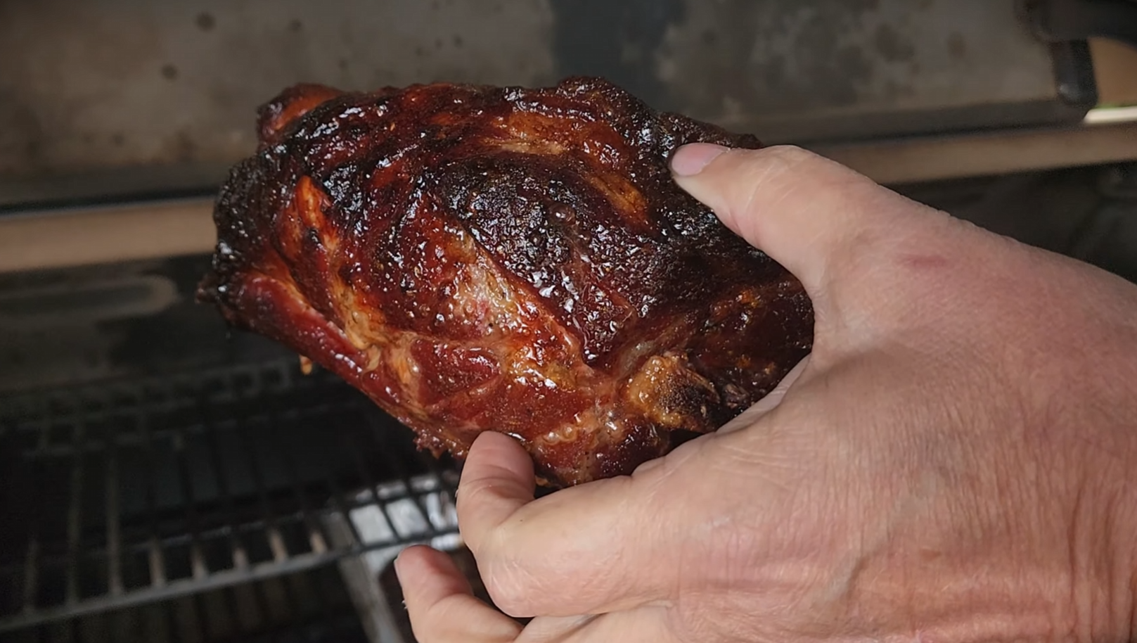 A close-up of hands holding a smoked, glistening piece of meat with a dark, caramelized crust, in front of a metal grill.