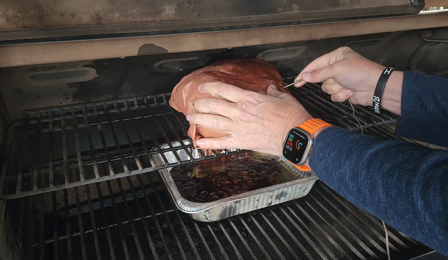 A person wearing a smartwatch checks the temperature of a large piece of meat on a grill, with a foil tray of liquid below to catch drippings.