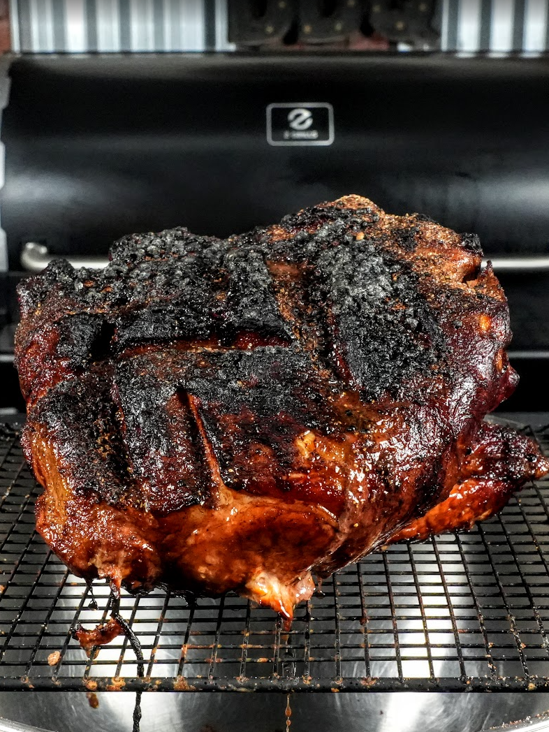 A large, charred, and caramelized piece of smoked meat rests on a wire rack in front of a grill. The meat has deep grill marks and a dark, crispy bark with glistening, juicy edges.