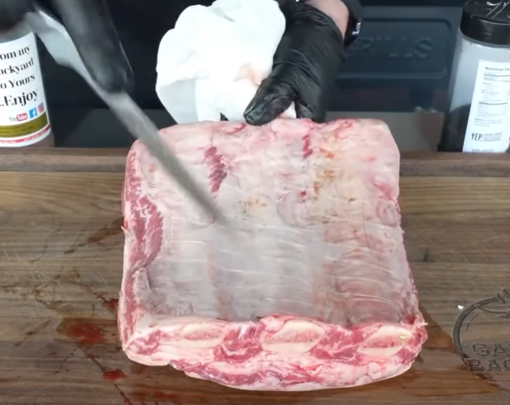 A person wearing black gloves uses a knife to remove the membrane from a rack of raw beef ribs on a wooden cutting board, with paper towels and seasonings nearby.