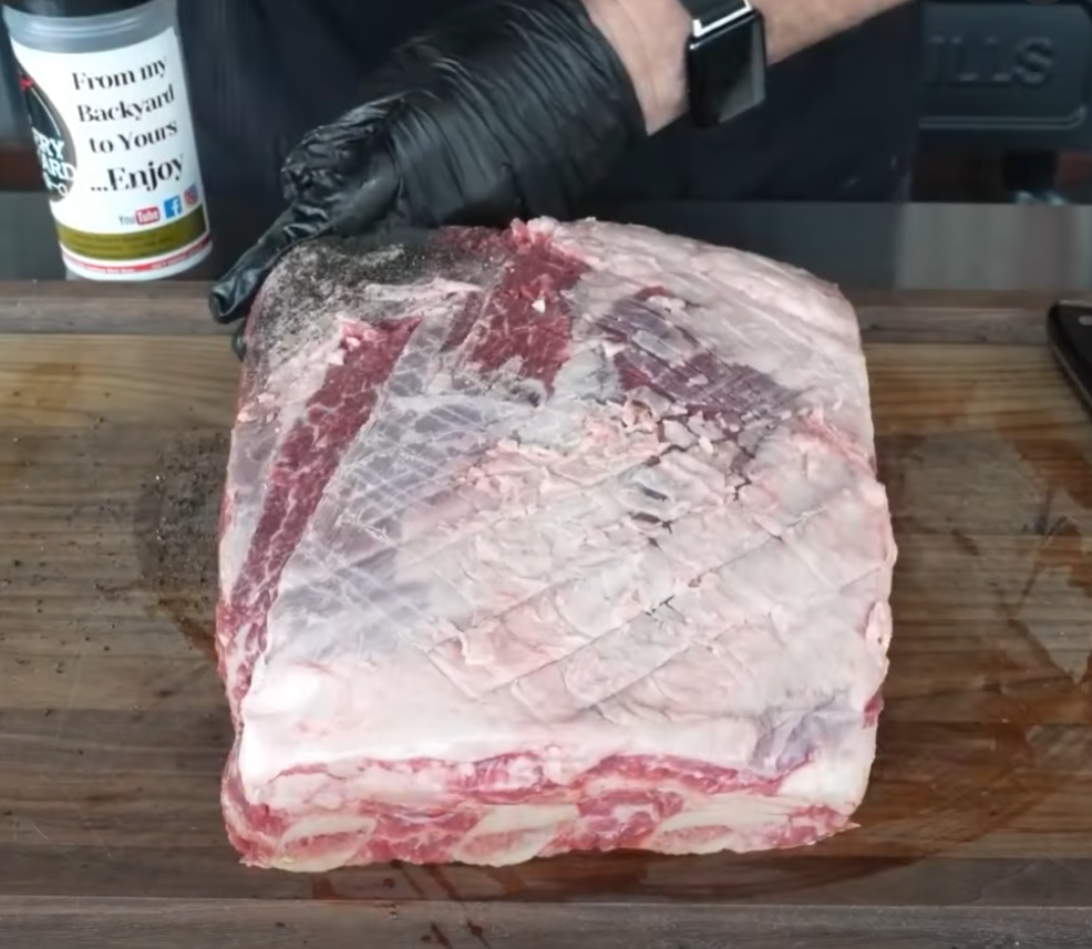 A person wearing a black glove seasons a large raw beef rib roast on a cutting board, with a seasoning container visible in the background.