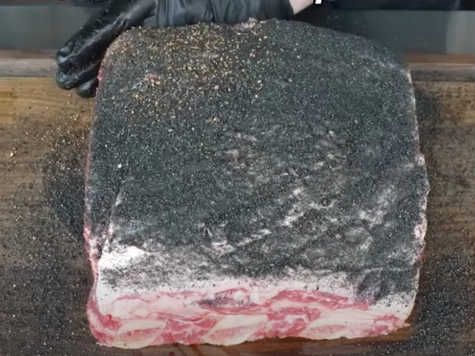 A large cut of raw beef brisket covered with a thick layer of black pepper and seasonings sits on a wooden board. A gloved hand holds the meat steady.