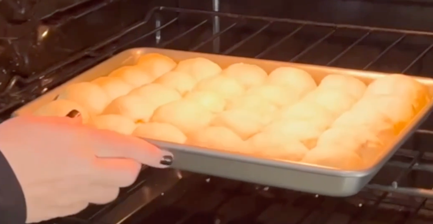 A hand with black nail polish places a baking tray of golden, round rolls onto an oven rack.