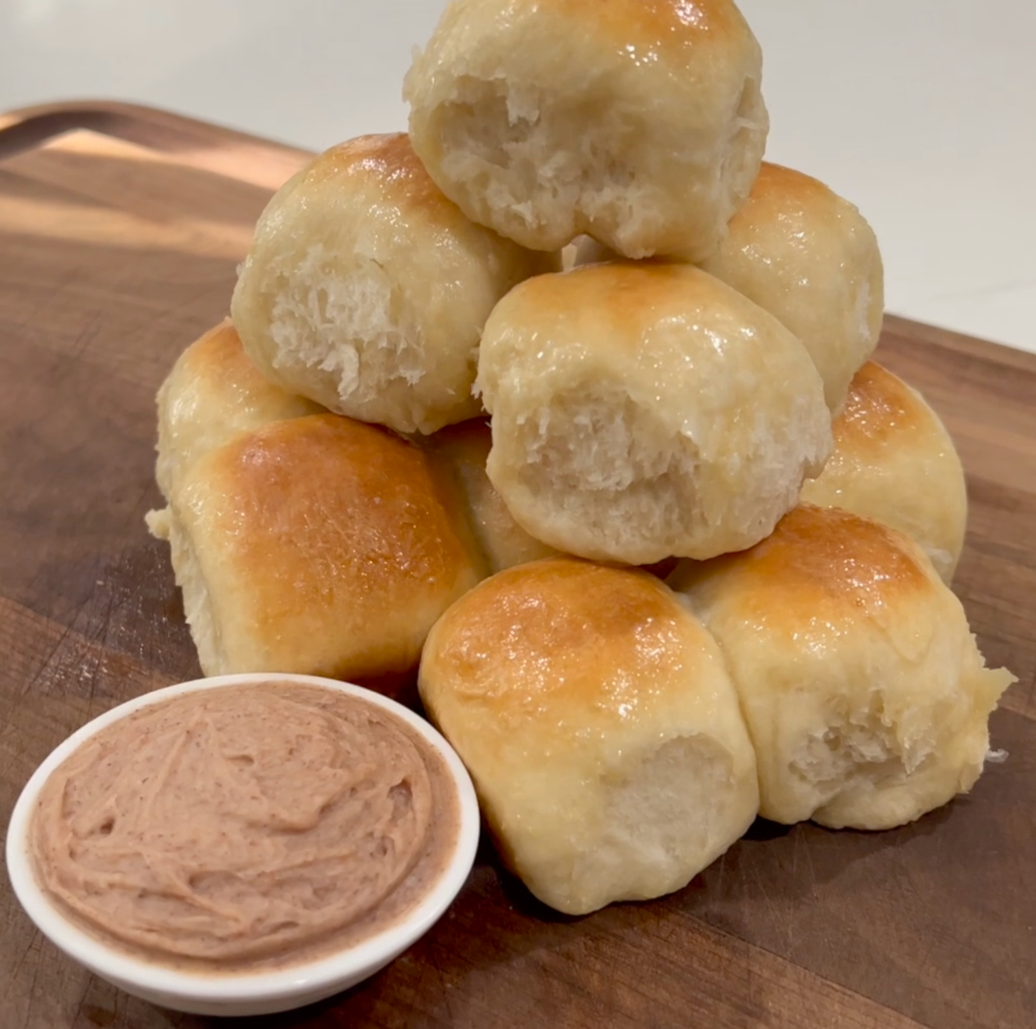 A stack of golden, fluffy dinner rolls sits on a wooden board next to a small white bowl filled with whipped cinnamon butter.