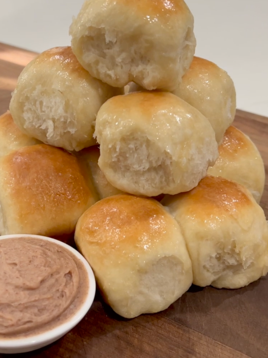 A pyramid stack of shiny, golden-brown dinner rolls sits on a wooden board next to a small bowl filled with light brown whipped butter.