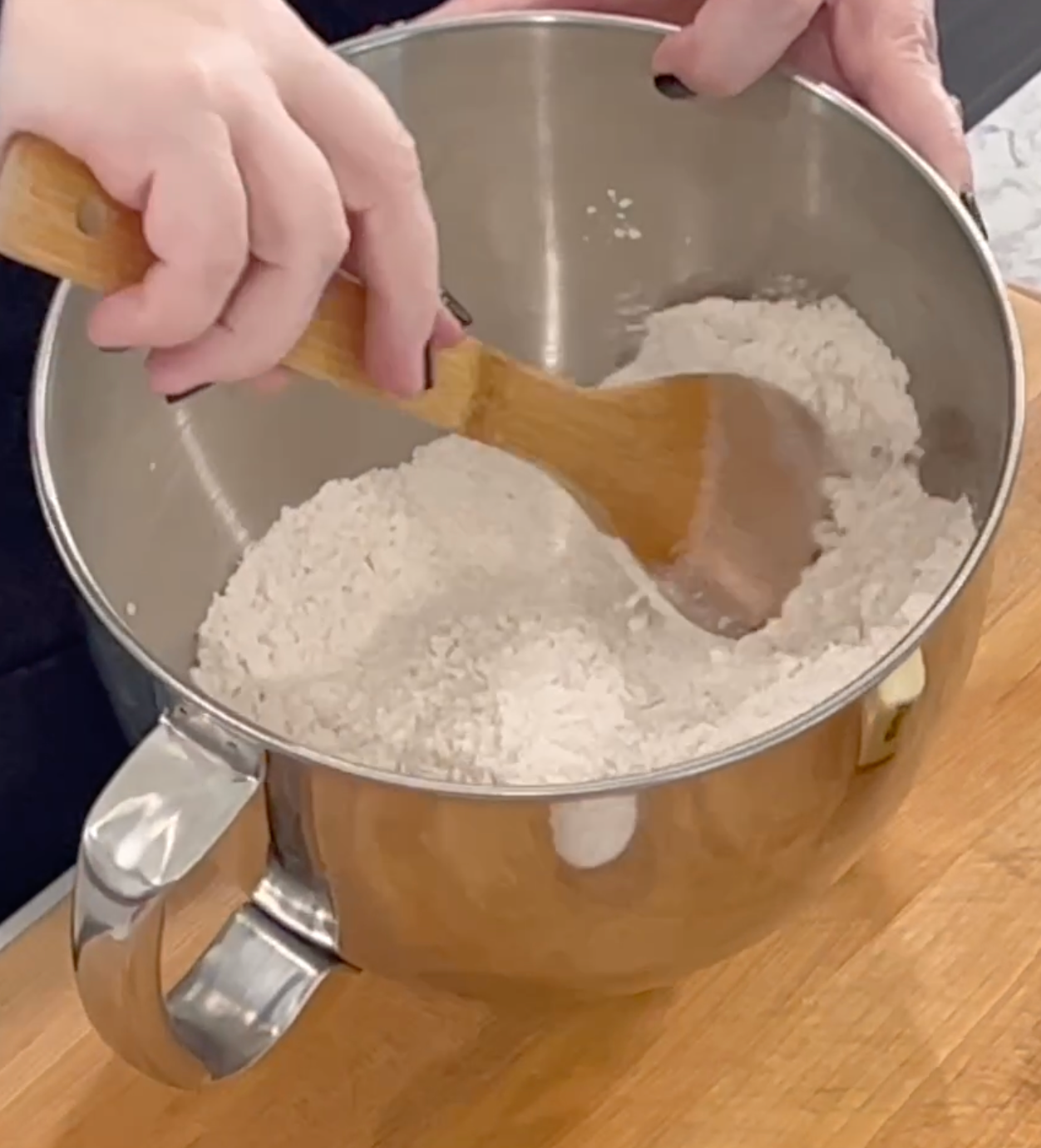 A person stirs flour in a metal mixing bowl with a wooden spoon on a wooden surface.