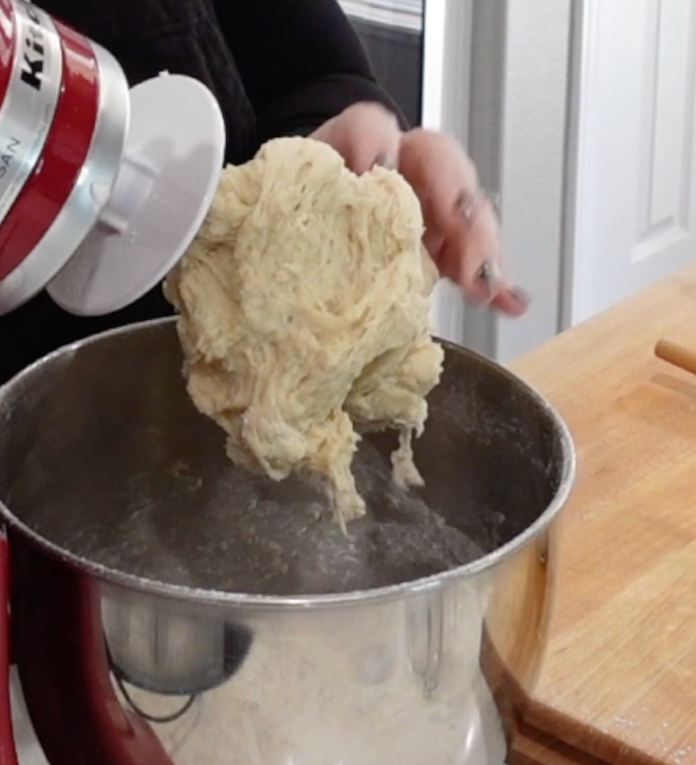 A person lifts bread dough from a stand mixer with a dough hook over a large metal mixing bowl on a wooden countertop.