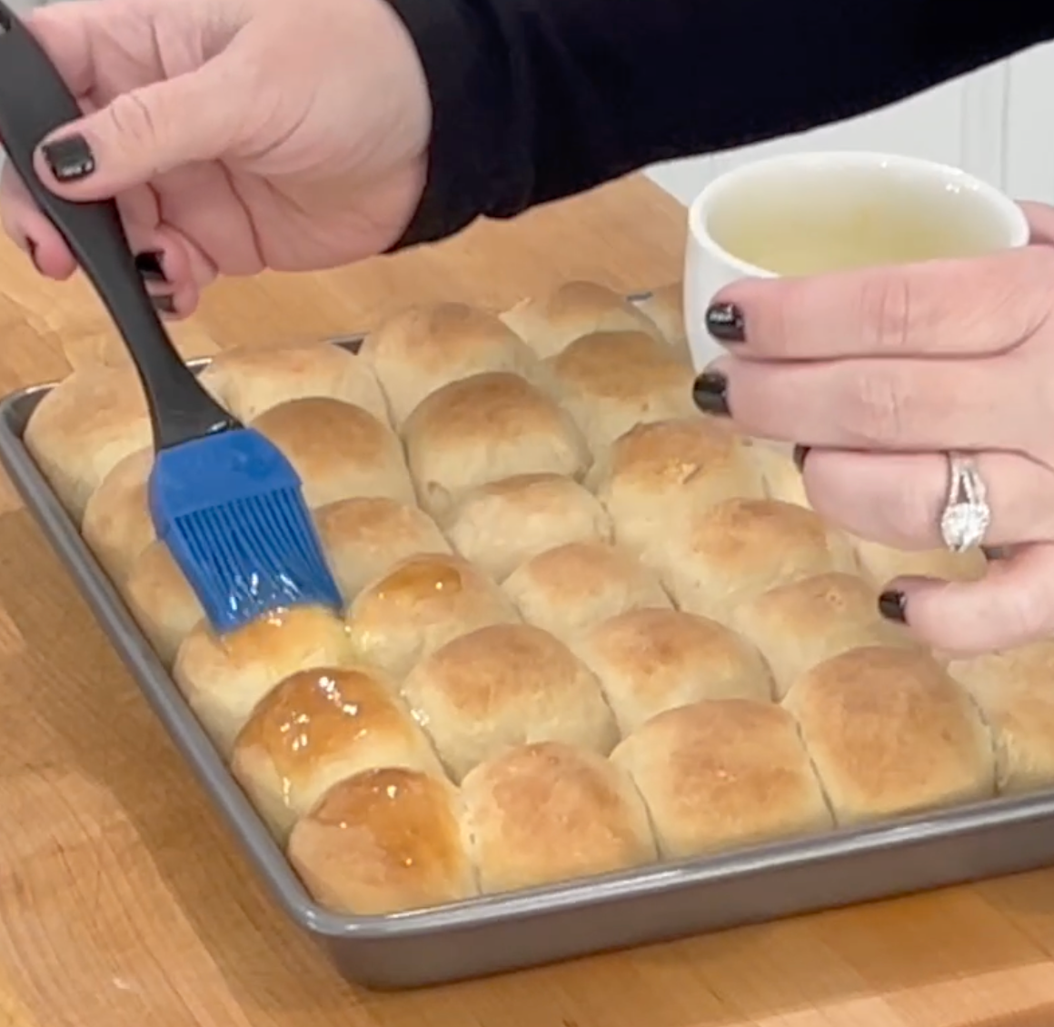 A person brushes melted butter onto freshly baked dinner rolls in a baking tray, using a blue pastry brush and holding a small white cup.