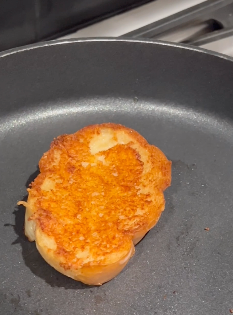 A slice of bread with a golden-brown, crispy crust is toasting in a nonstick frying pan on a stove.