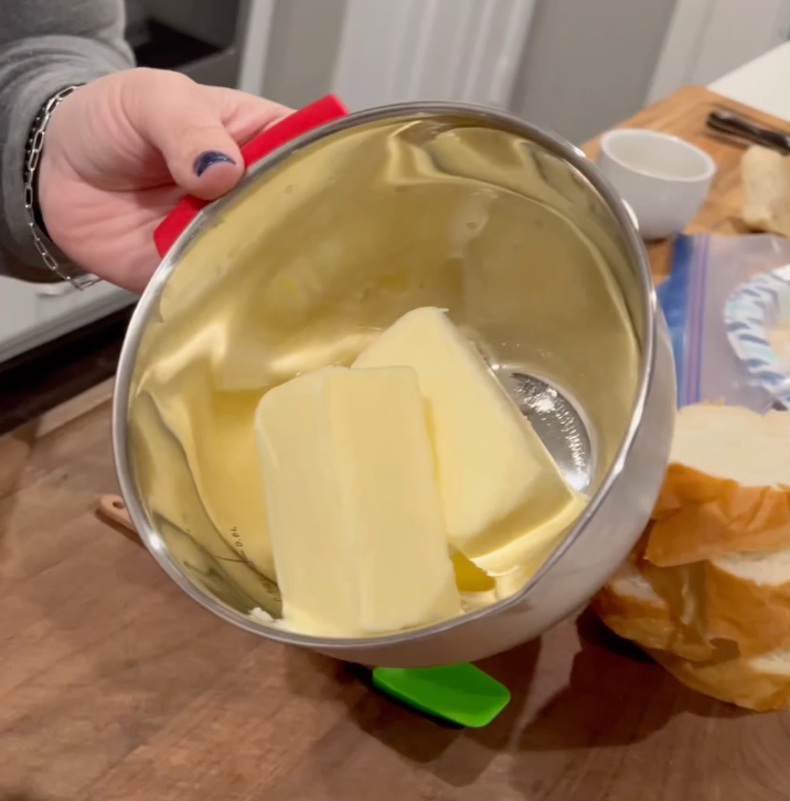 A hand holds a metal mixing bowl with two sticks of butter inside, above a wooden countertop with sliced bread and kitchen items nearby.