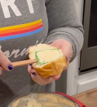 A person spreads green icing on a cream-filled pastry using a wooden stick. They wear a gray shirt with colorful stripes and text.