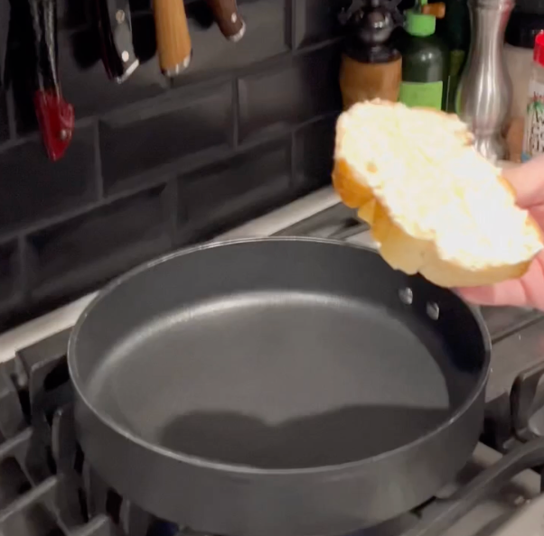 A hand holds a piece of sliced bread over a black frying pan on a stovetop, with kitchen utensils and condiments visible in the background.