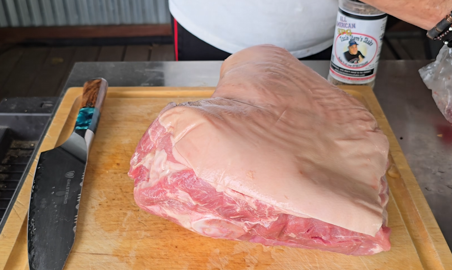 A large raw pork roast sits on a wooden cutting board next to a knife. A person holding a can stands in the background, partially visible.