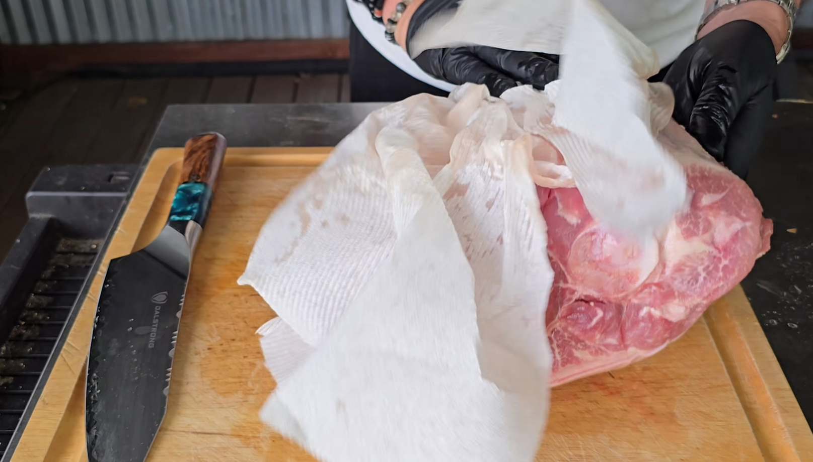 A person wearing black gloves pats a large piece of raw meat dry with paper towels on a wooden cutting board beside a knife with a colorful handle.